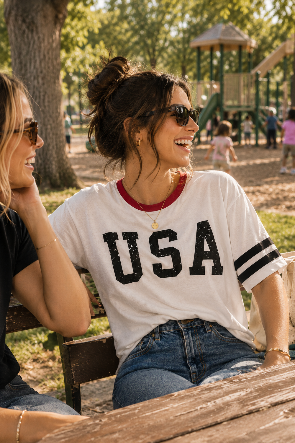 A woman sitting outdoors at a park bench wearing a white short-sleeve t-shirt with 'USA' printed in black across the chest and blue jeans. Another person is partially visible on the left.